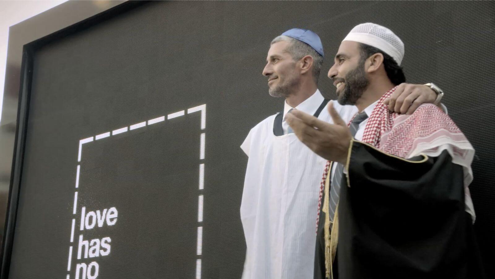 Jewish man with arm around Muslim man next to digital sign that reads 