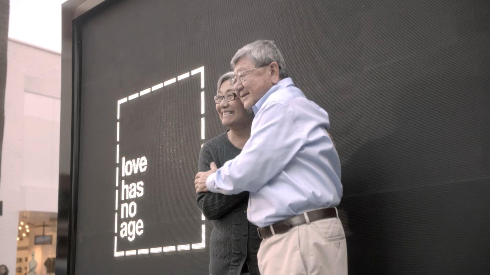 Older couple smiling and hugging next to digital sign that reads 