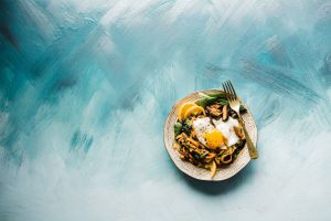 A plate with spinach, mushrooms and noodles cooked together with a fried egg on top is sitting on a blue painted table