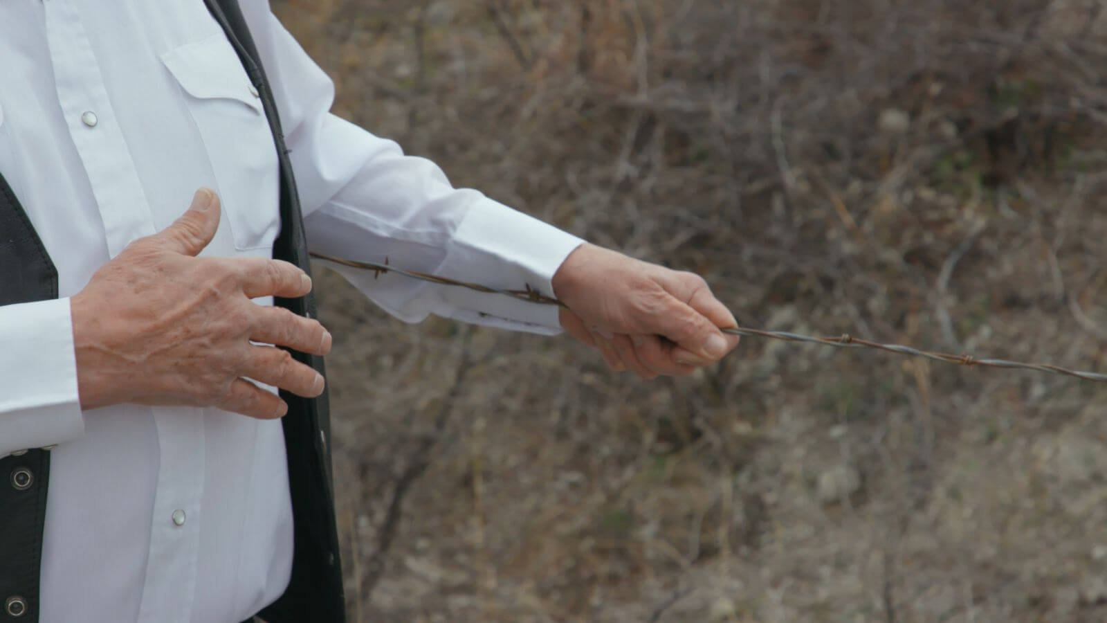 Hands of old man in white shirt and black vest touching a barbed wire fence