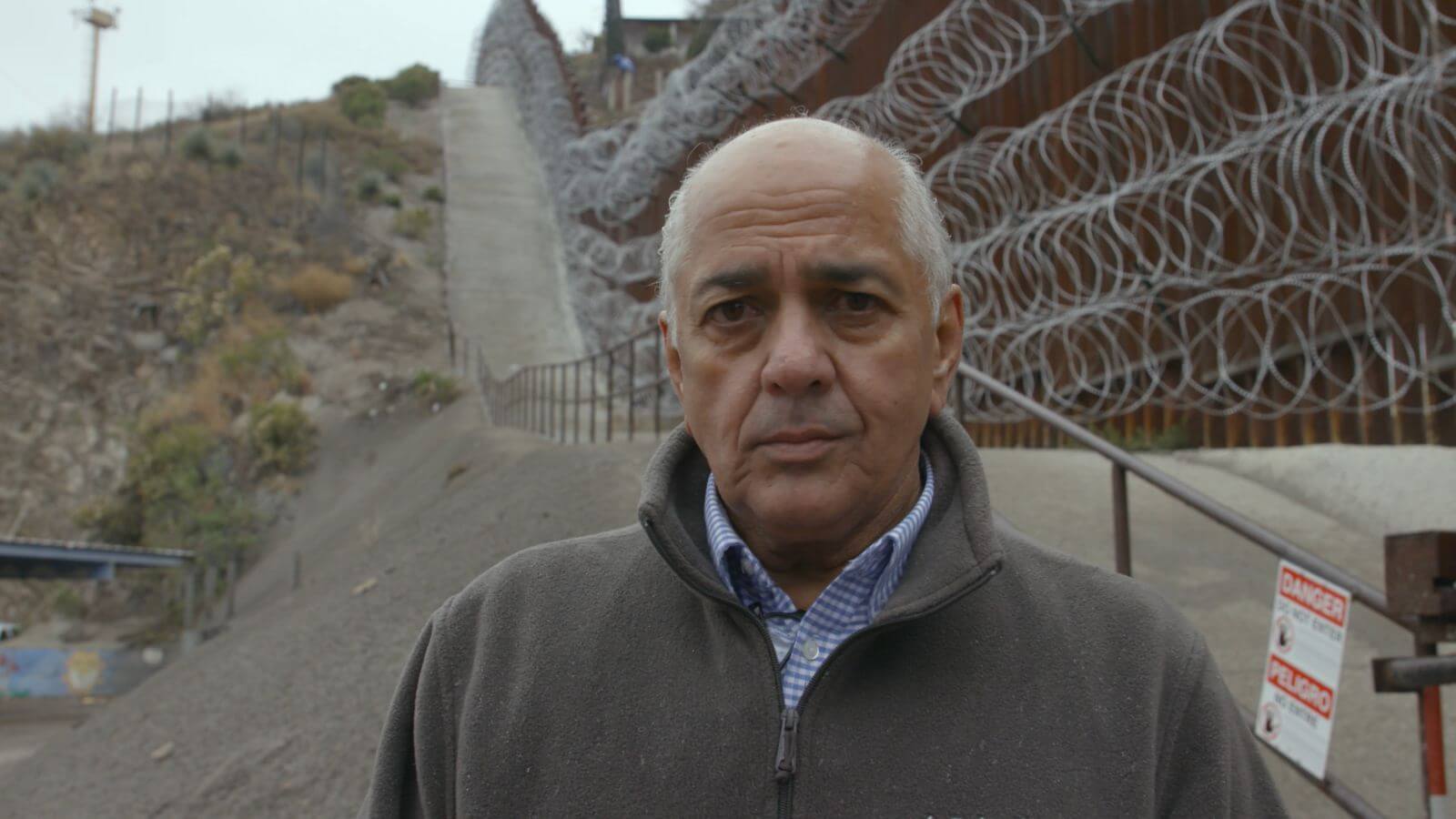 Older man standing in front of a large border fence with long, running loops of barbed wire