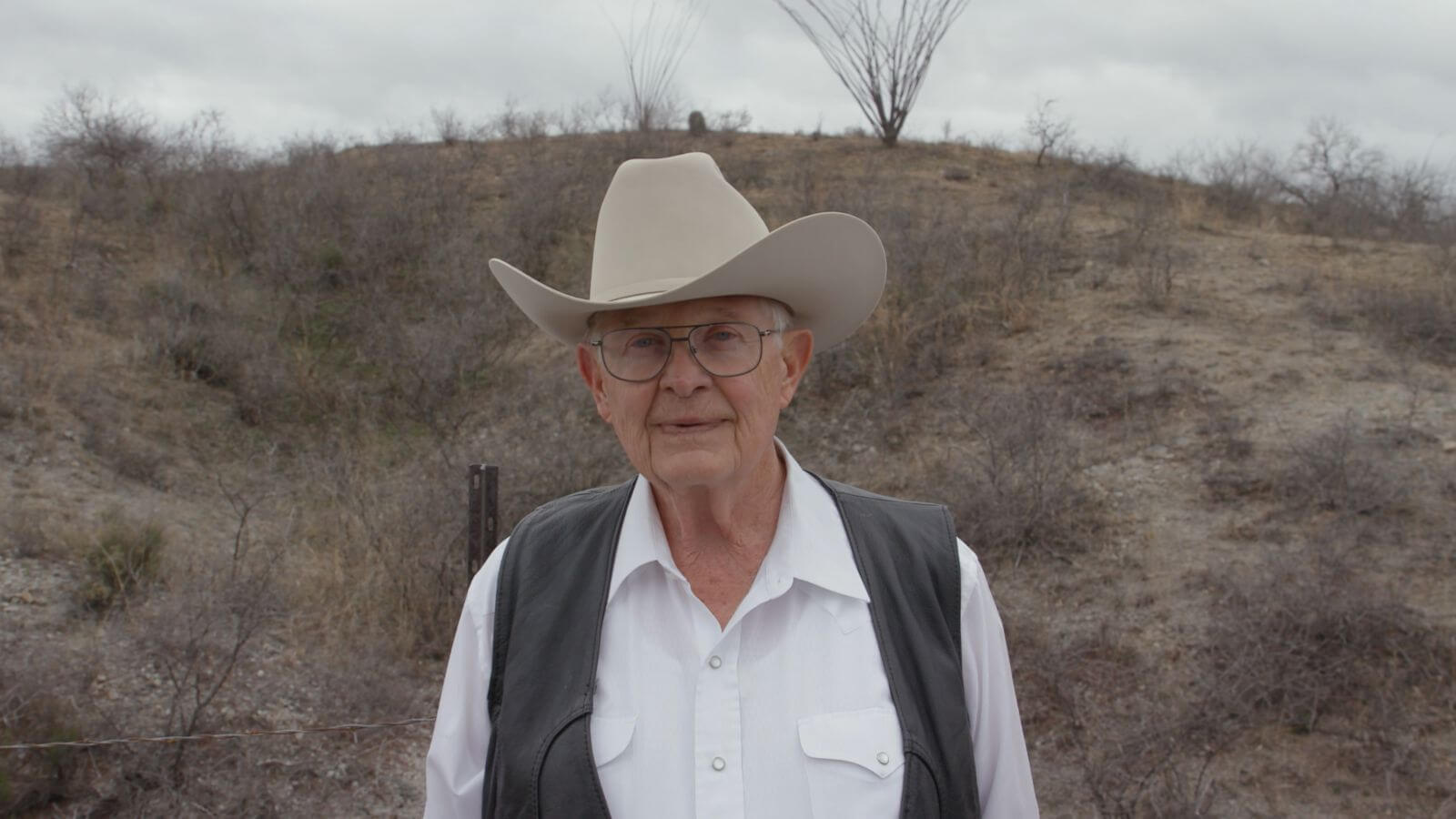 Old man in a white shirt, black vest and tan cowboy hat outside in a plains/desert area