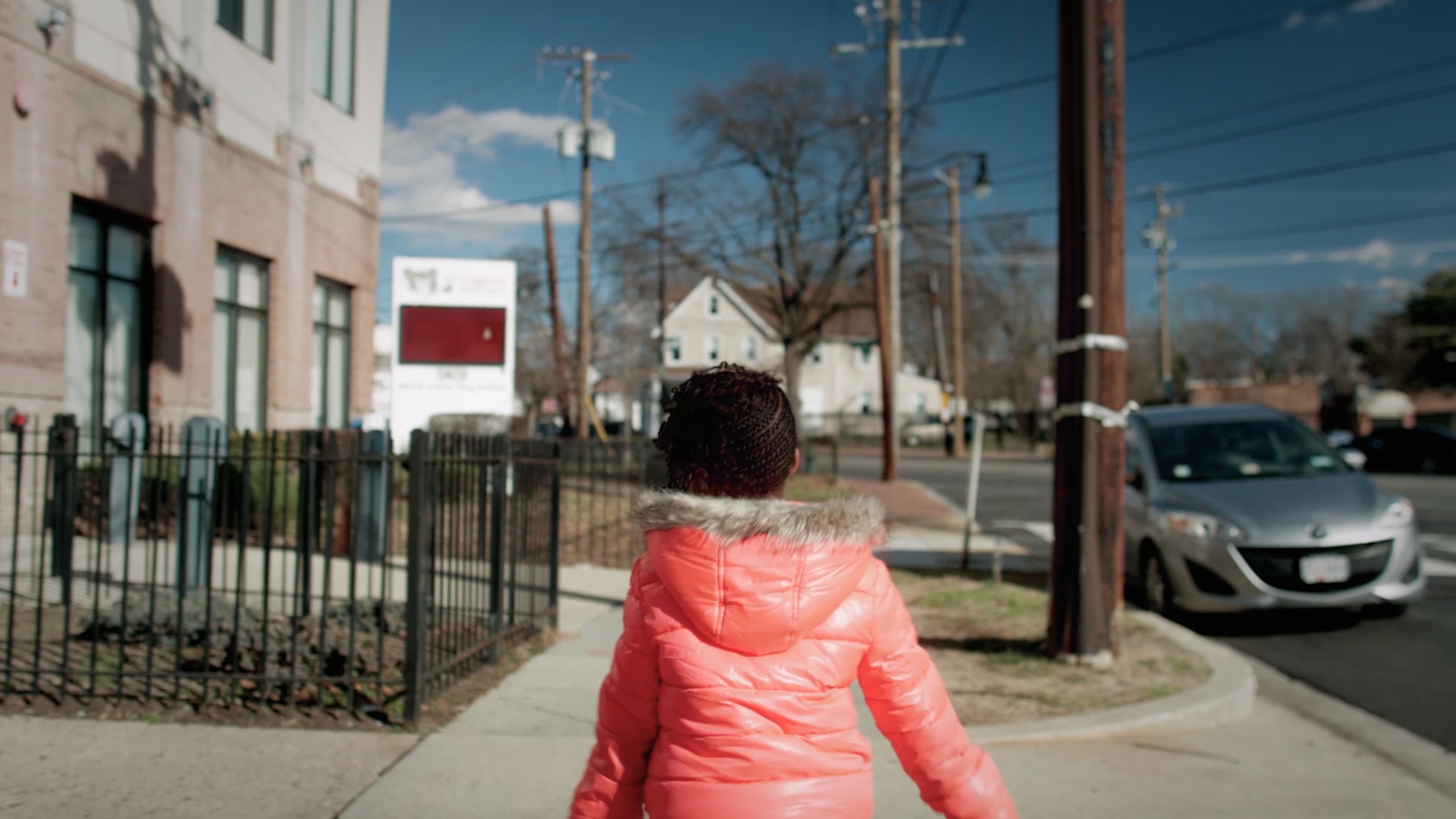 A young girl in a pink puffy winter coat walking away down a city sidewalk
