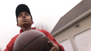 Boy wearing a ball cap and red coat holding a basketball and standing in front of a two car garage