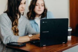 Two women sitting at a wooden table looking at the screen of a Samsung Chromebook