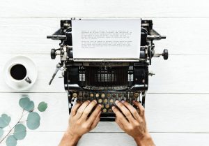Top down view of man's hands on a very old black typewriter with a cup of black coffee sitting next to it on the left
