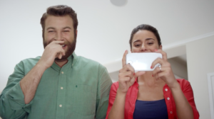Bearded man in green oxford smiling and standing next to woman taking a picture with a white cell phone