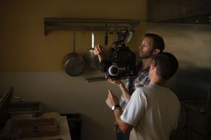 Two men with a video camera standing in a kitchen discussing the shot they are taking