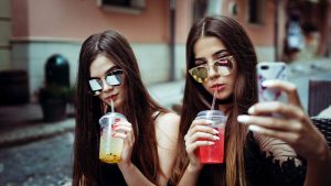 Two girls with long brown hair and sunglasses sipping cold drinks while one girl takes a selfie of them together